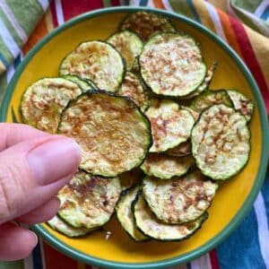 Finger holding a zucchini chip cooked in the microwave over a yellow plate with more zucchini chips.