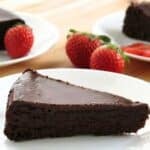 Flourless chocolate cake with ganache on a white plate with strawberries and more slices of cake behind it.