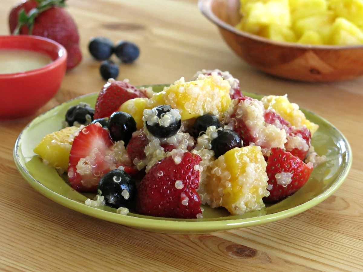 Serving of quinoa fruit salad on a plate with honey lime dressing and fruit in the backgroun.