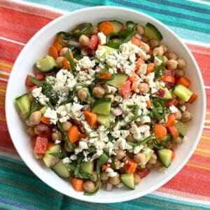 Chickpea feta cucumber salad with red onions, carrots, and tomatoes in a white bowl on a turquiose and orange cloth.