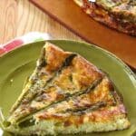 Slice of crustless asparagus quiche on a plate with part of a full quiche showing behind it.