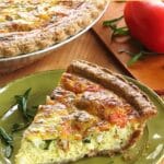 Slice of bacon tomato quiche on a plate with chopped basil and the full pie behind it.