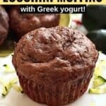 A healthy chocolate zucchini muffin made with Greek yogurt and chocolate chips on small cake pedestal surrounding by shreds with zucchini and more muffins in the background.