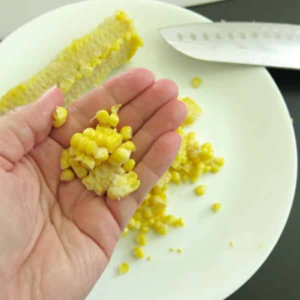 Hand holding previously frozen corn kernels with a cob and knife on a cutting board