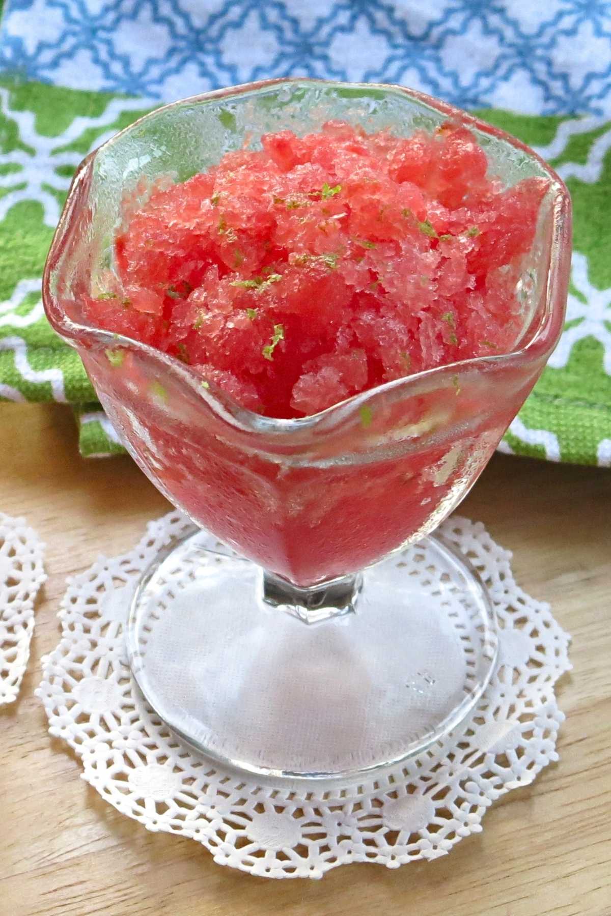 Watermelon granita with lime zest on top in a glass serving cup on a white doily.