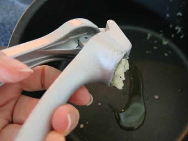 Hand squeezing garlic press over pan with oil