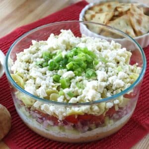 7-layer Greek Dip in a glass bowl with layers of hummus, tsatziki, tomatoes, red onions, feta cheese, and green onions.