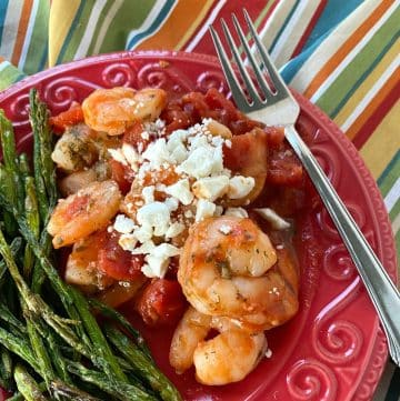 Serving of Greek shrimp with tomatoes and feta cheese on plate next to asparagus.
