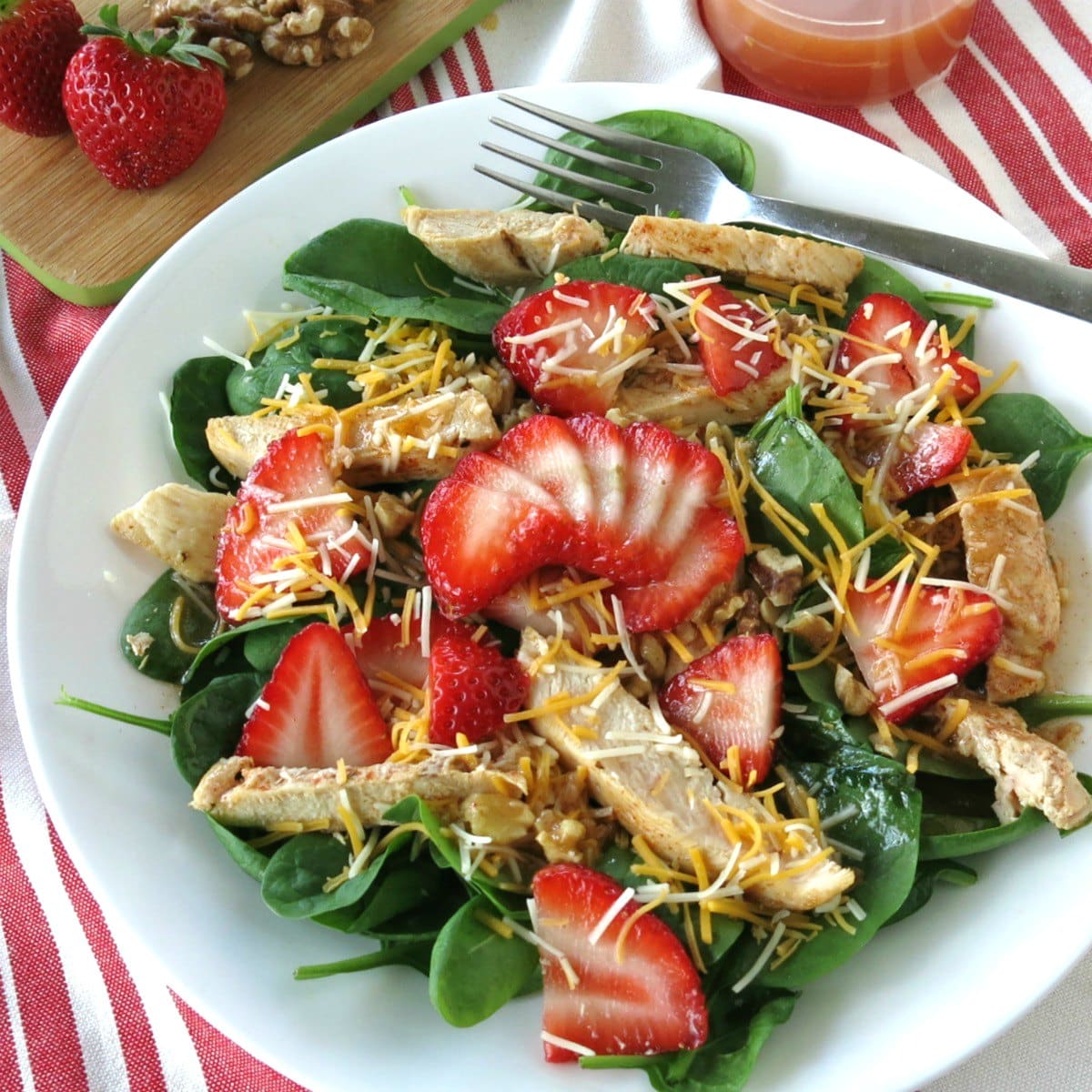 Spinach Strawberry Walnut Salad topped with chicken on a plate with walnuts and dressing in the background.