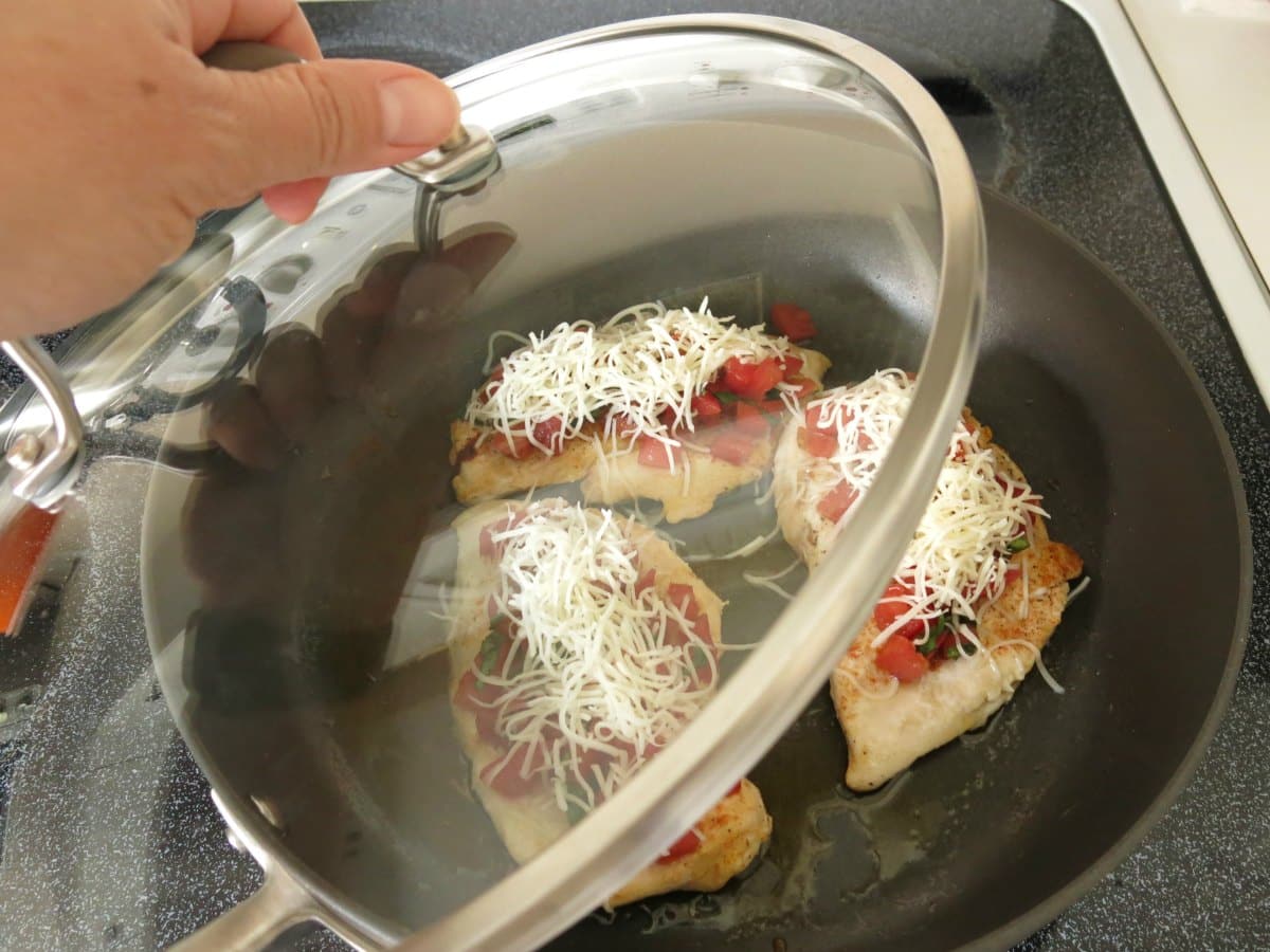 Glass lid being placed on skillet with chicken topped with bruschetta and cheese inside.