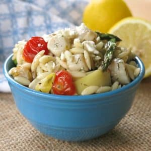 Warm orzo salad with scallops, artichokes, tomatoes, asparagus, and feta in a bowl with lemons behind it.