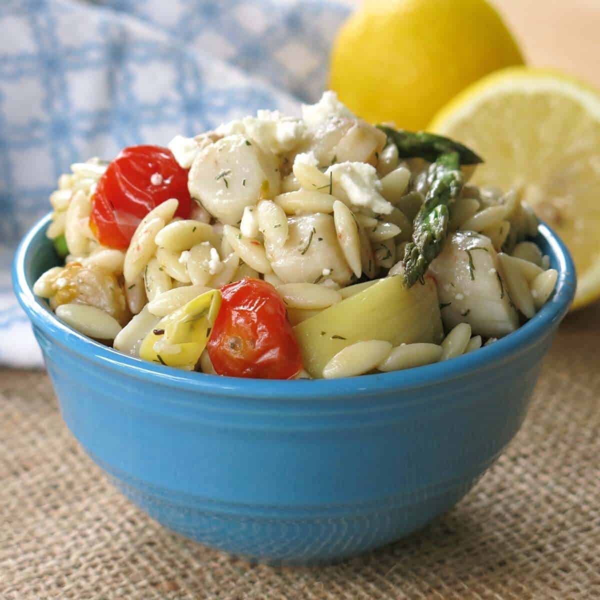 Warm orzo salad with scallops, artichokes, tomatoes, asparagus, and feta in a bowl with lemons behind it.