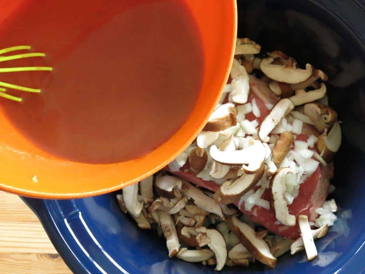 Uncooked beef shoulder, mushooms, and onions in crock-pot and a bowl with sauce being pouring into it.