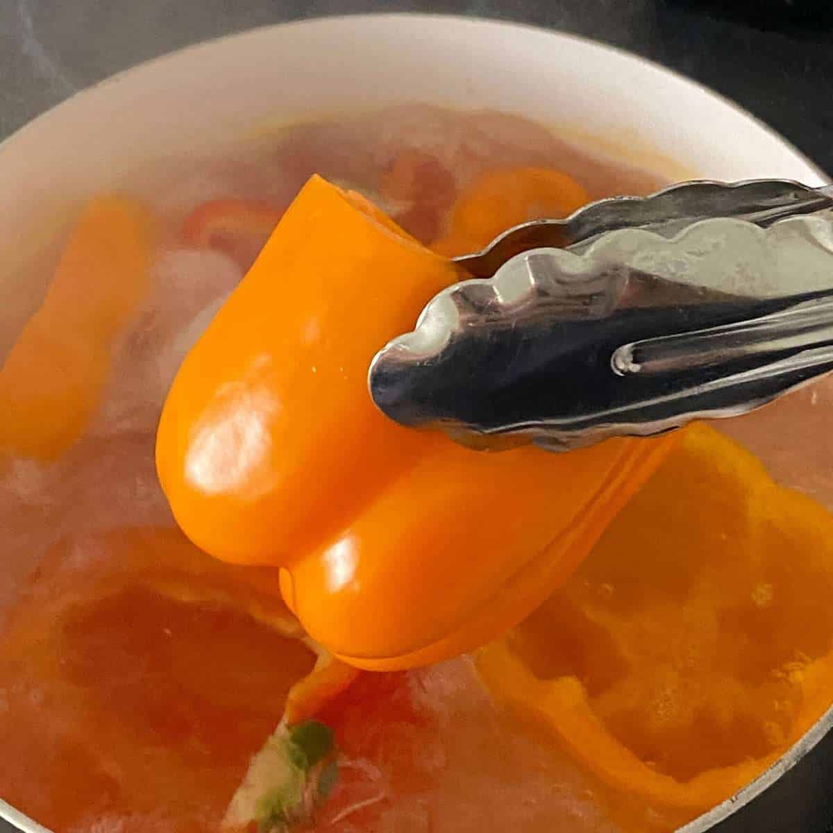 Tongs pulling an orange bell pepper out of a pot of boiling water.