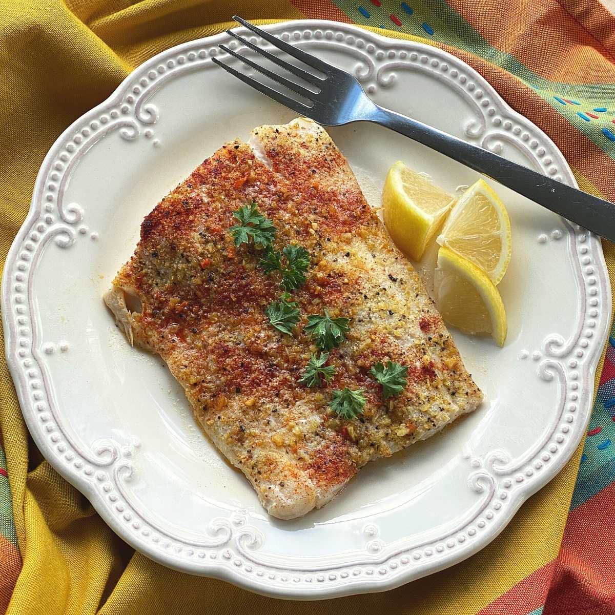 Filet de corvina cuit au four avec une croûte au poivre citronné sur une assiette avec des quartiers de citron et une fourchette.