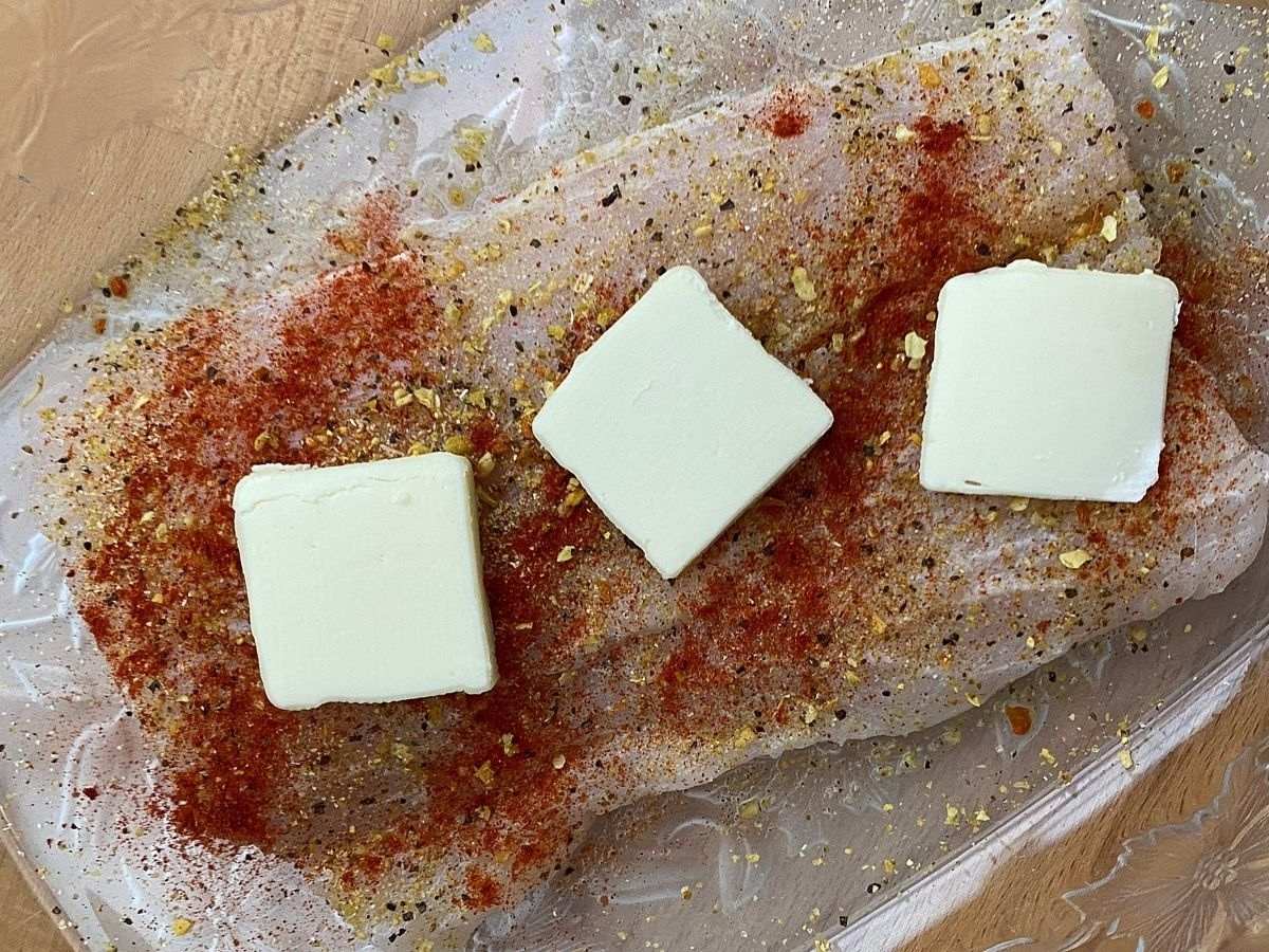 Uncooked corvina fillet with seasonings and butter in baking dish.