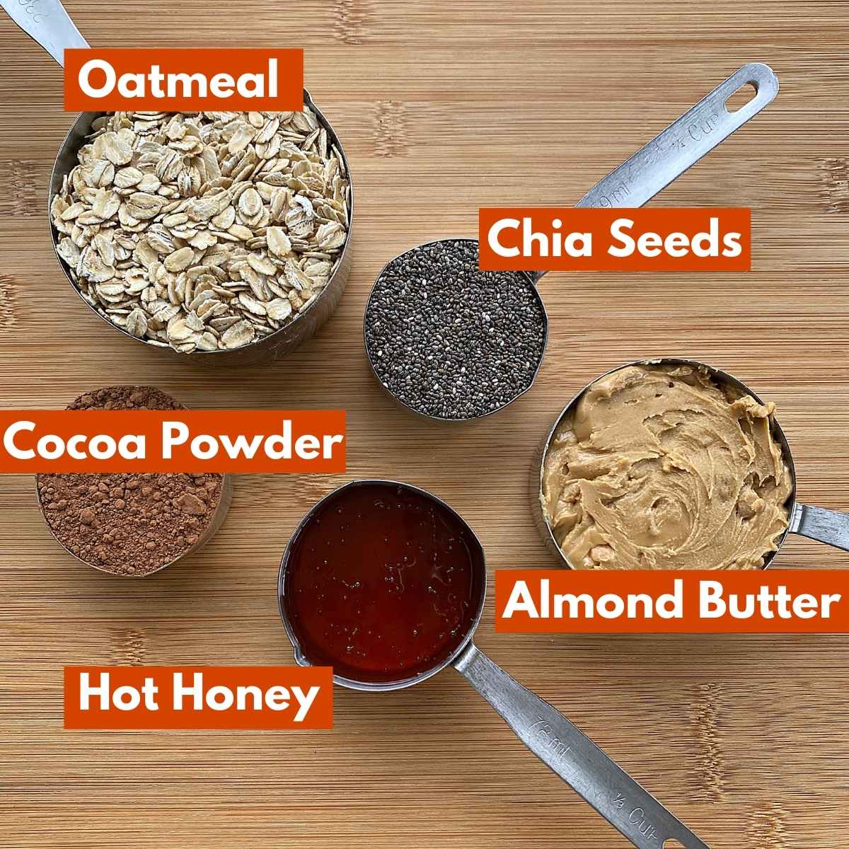 5 Ingredients in measuring cups with labels: oatmeal, chia seeds, cocoa powder, almond butter, hot honey.