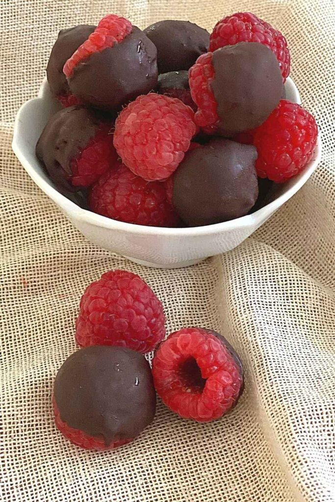 3 raspberries on a napkin with bowl of dark chocolate raspberries and a few plain ones in it.