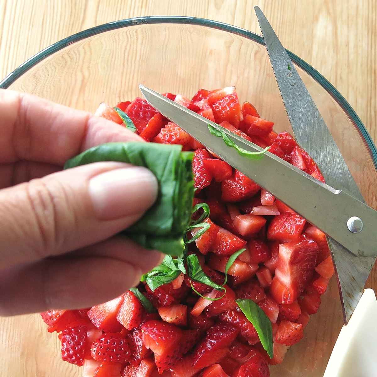 Hand holding basil that is stacked and rolled and scissors that cut it into strips over bowl of diced strawberries.