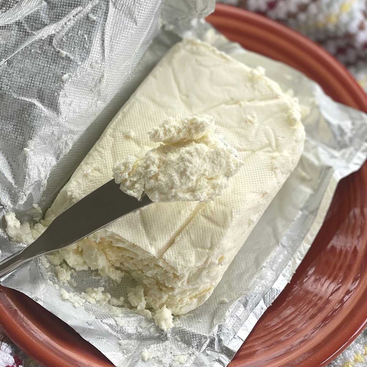 Knife with thawed cream cheese on it over a block of cream cheese in an open wrapper.
