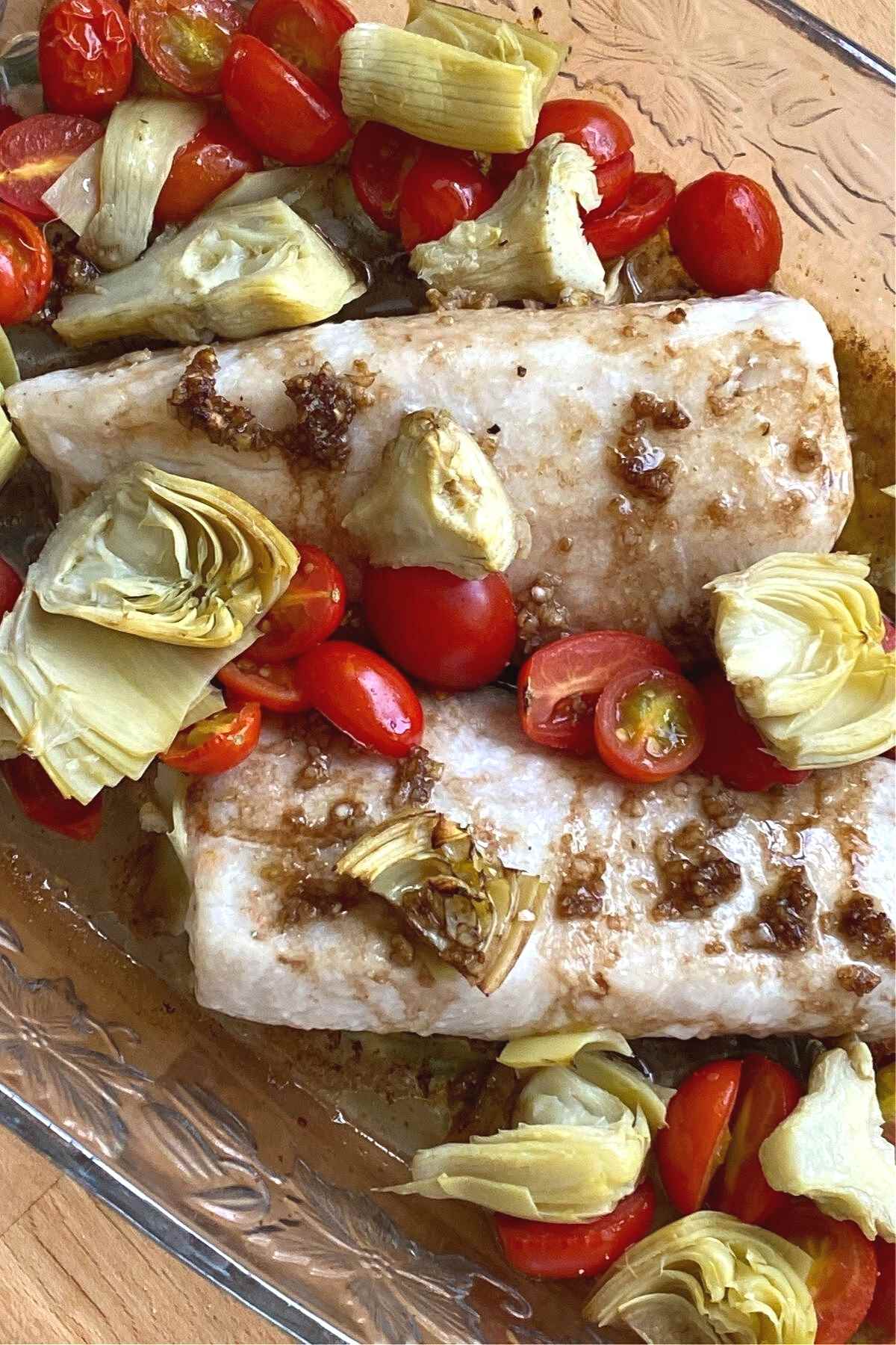 Cooked Cobia fish fillets with artichokes and tomatoes in a baking dish.