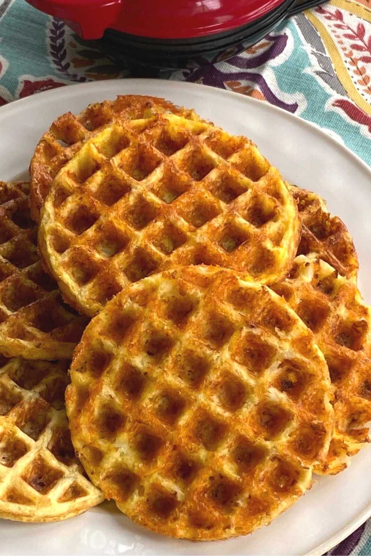 Cauliflower chaffles scattered on plate with Dash waffle iron behind it.