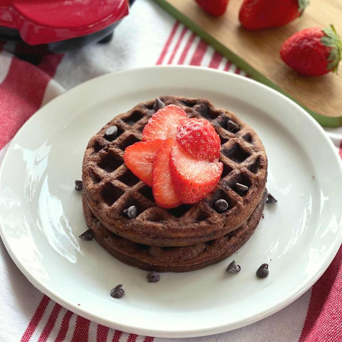 2 chocolate chaffles on a plate with strawberries and chocolate chips on top and a Dash waffle iron behind them.