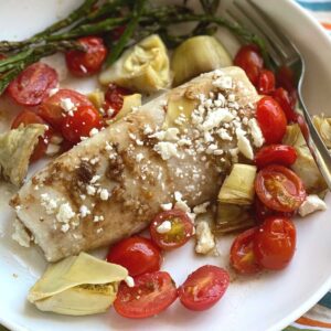 Mediterranean Baked Fish recipe on plate with tomatoes, artichokes and feta cheese.
