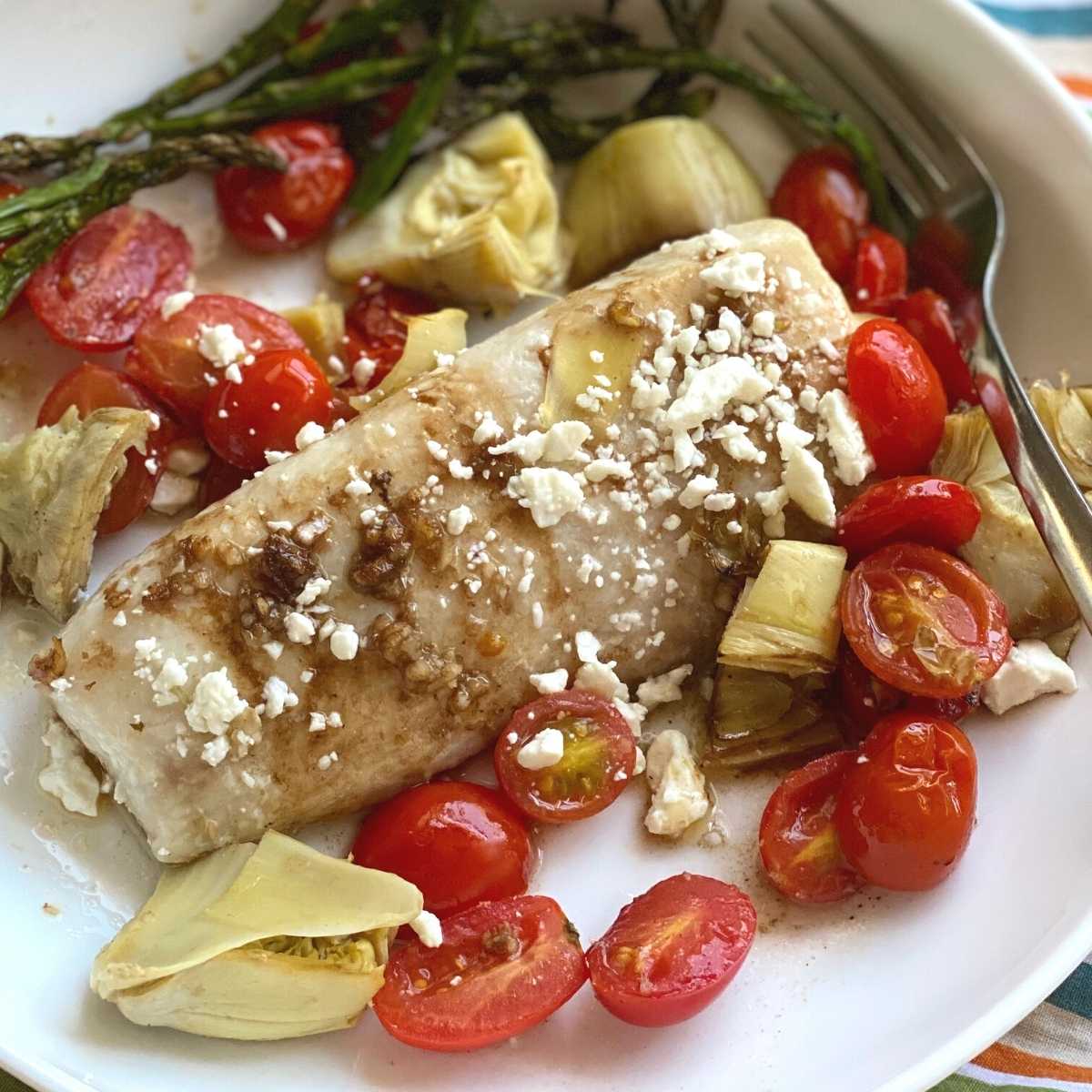 Mediterranean Baked Fish recipe on a plate with tomatoes, artichokes and feta cheese.