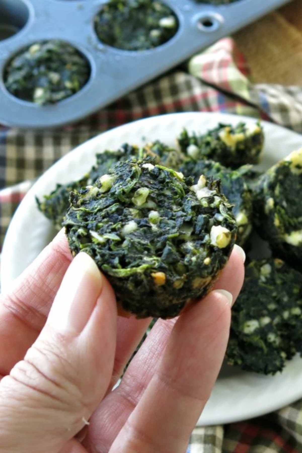 Hand holding one keto spinach feta egg cup with a pan of additional muffins behind it.