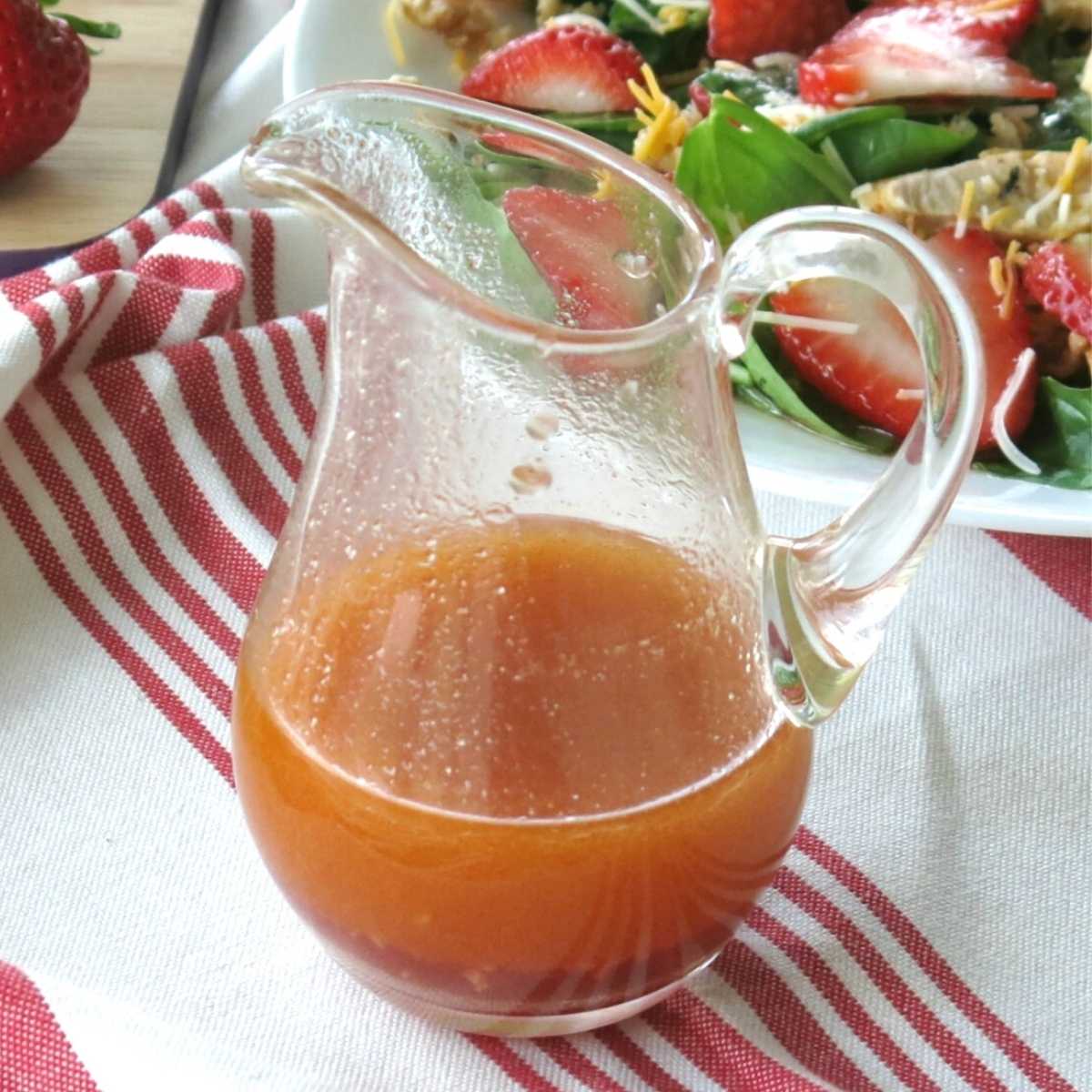 Sweet red wine vinegar dressing in a serving jar with strawberry salad behind it.