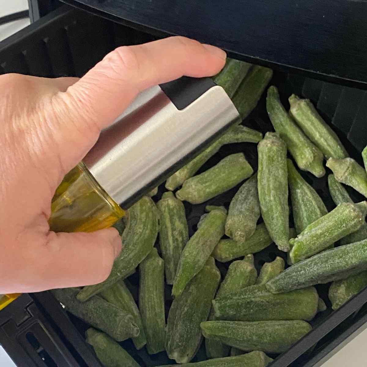 Hand holding olive oil mister over okra pods in an air fryer.