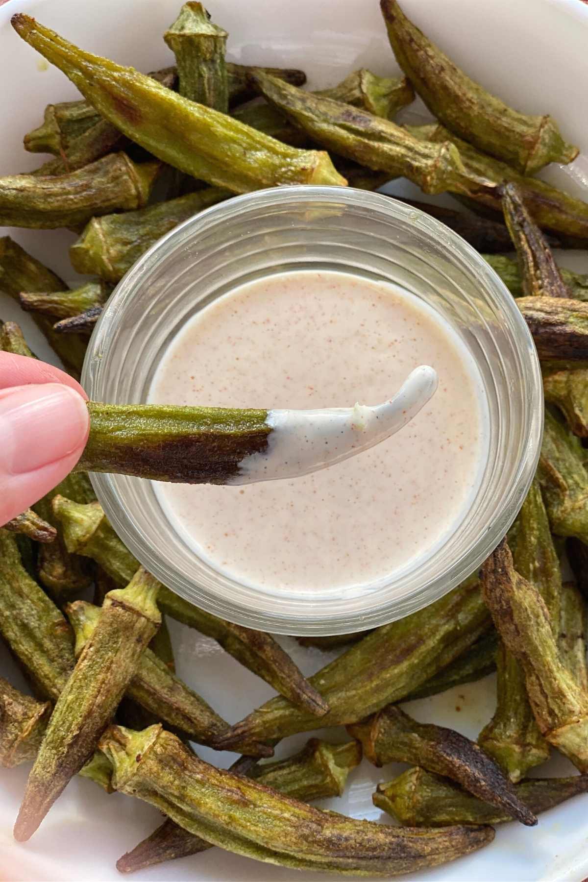 Fingers holding an okra fry dipped in aioli over a plate of okra fries.