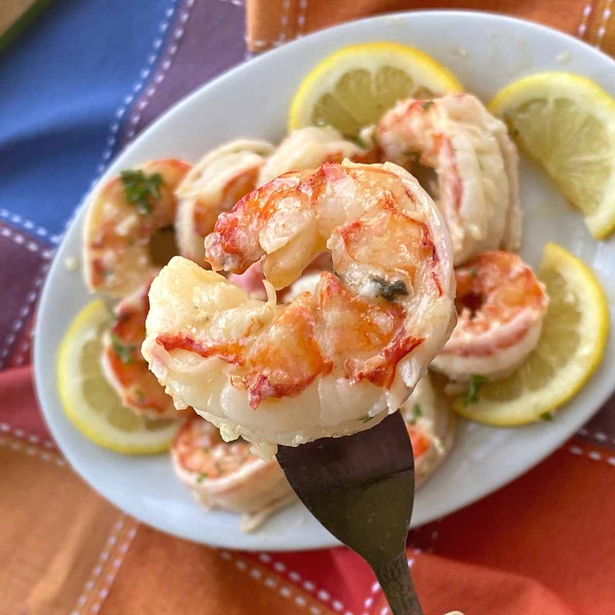 Fork holding up a cooked red Argentine shrimp covered in scampi sauce with more shrimp on the plate below.