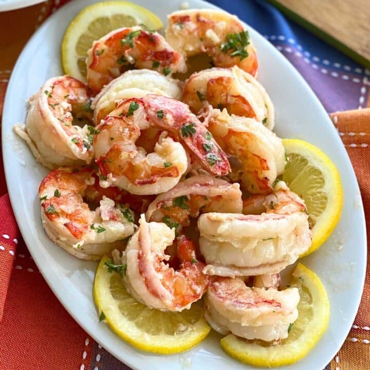 Argentine shrimp in a lemon, butter, garlic, scampi sauce on a white platter surrounded by slices of lemon.