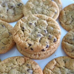 Eggless Chocolate Chip Cookies on a doily with plate of cookies and glass of milk behind it.