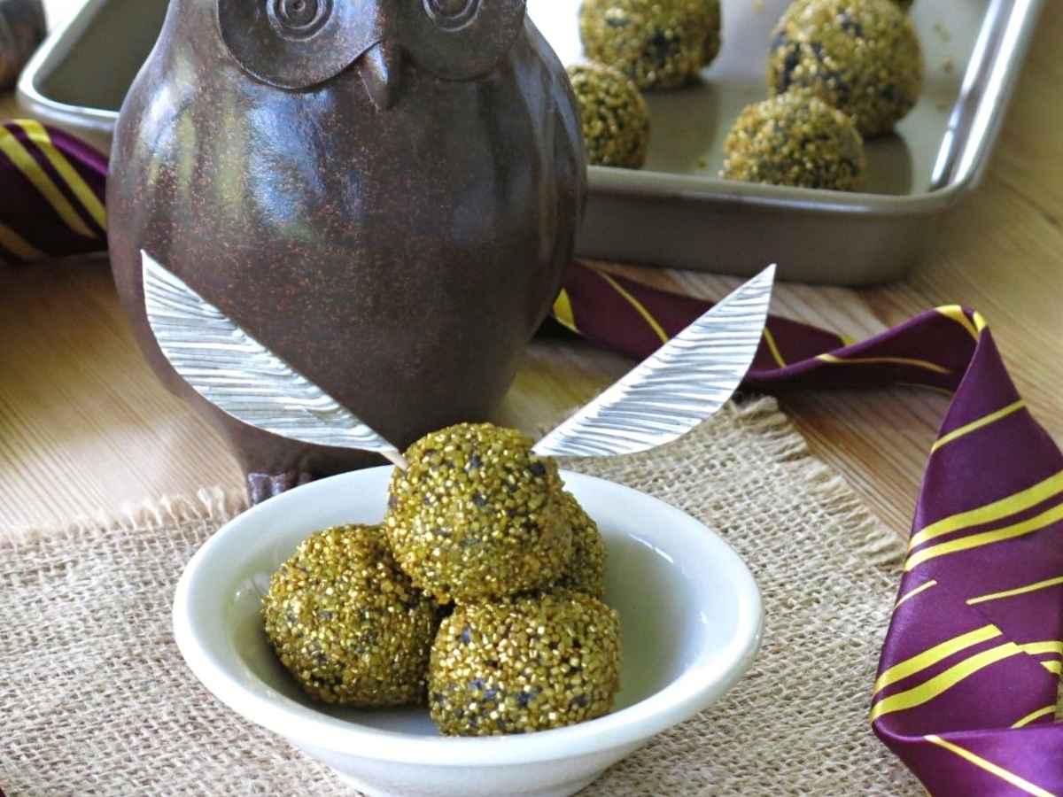 Vegan Golden snitch truffles in a bowl with an owl, Harry Potter neck tie, and pan of more balls behind it.