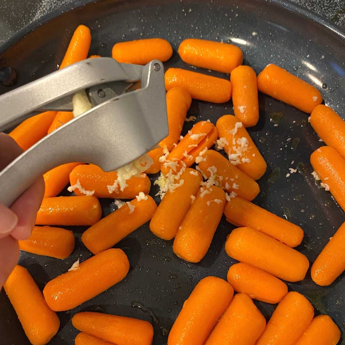 Garlic being added to a skillet of baby carrots with garlic press.