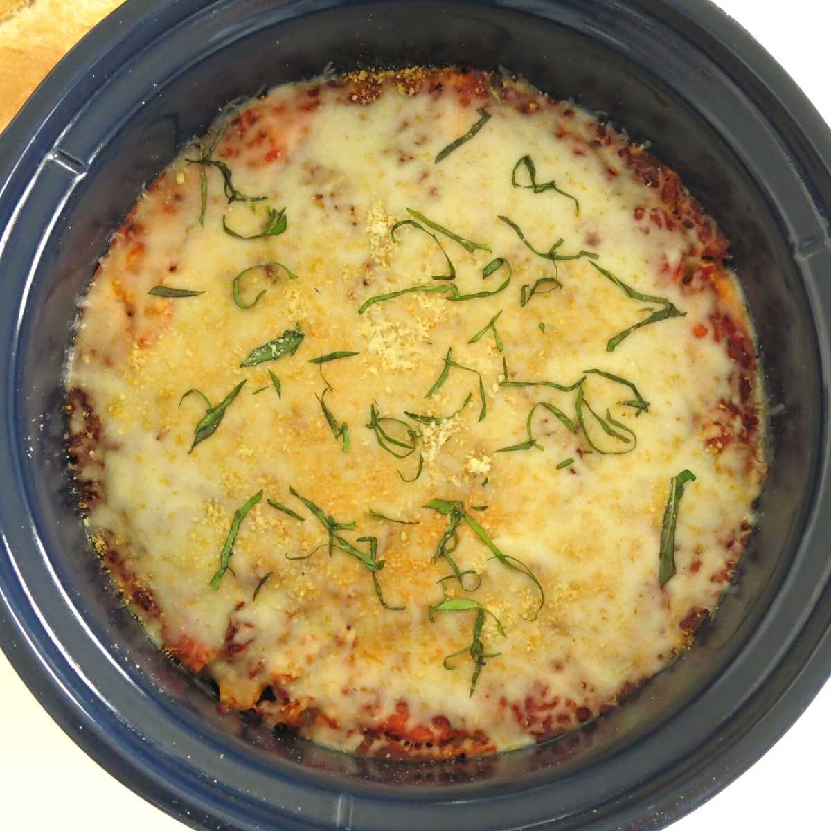 Overhead shot of baked ziti in a crock-pot with cheese and basil on top.