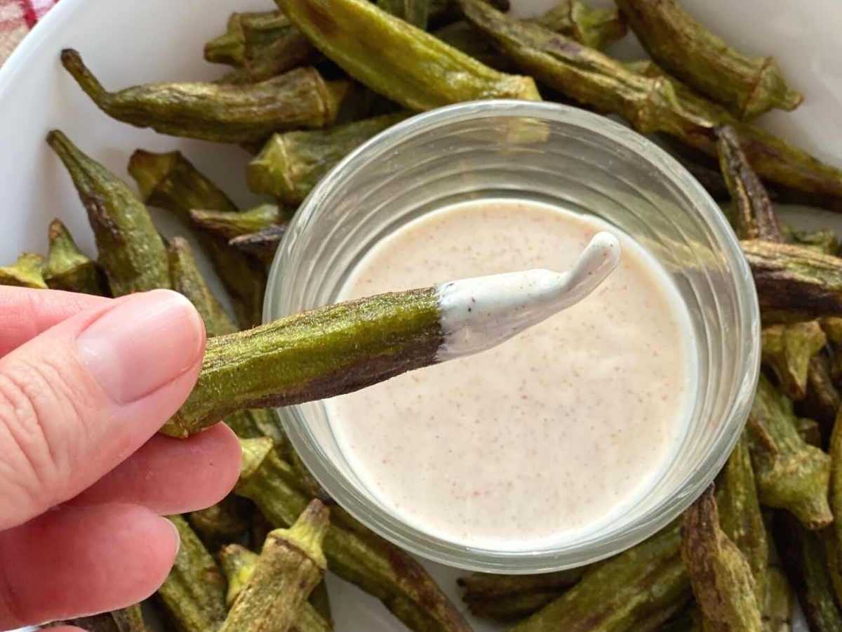 Fingers holding an okra fry over a plate with okra fries an dipping sauce.