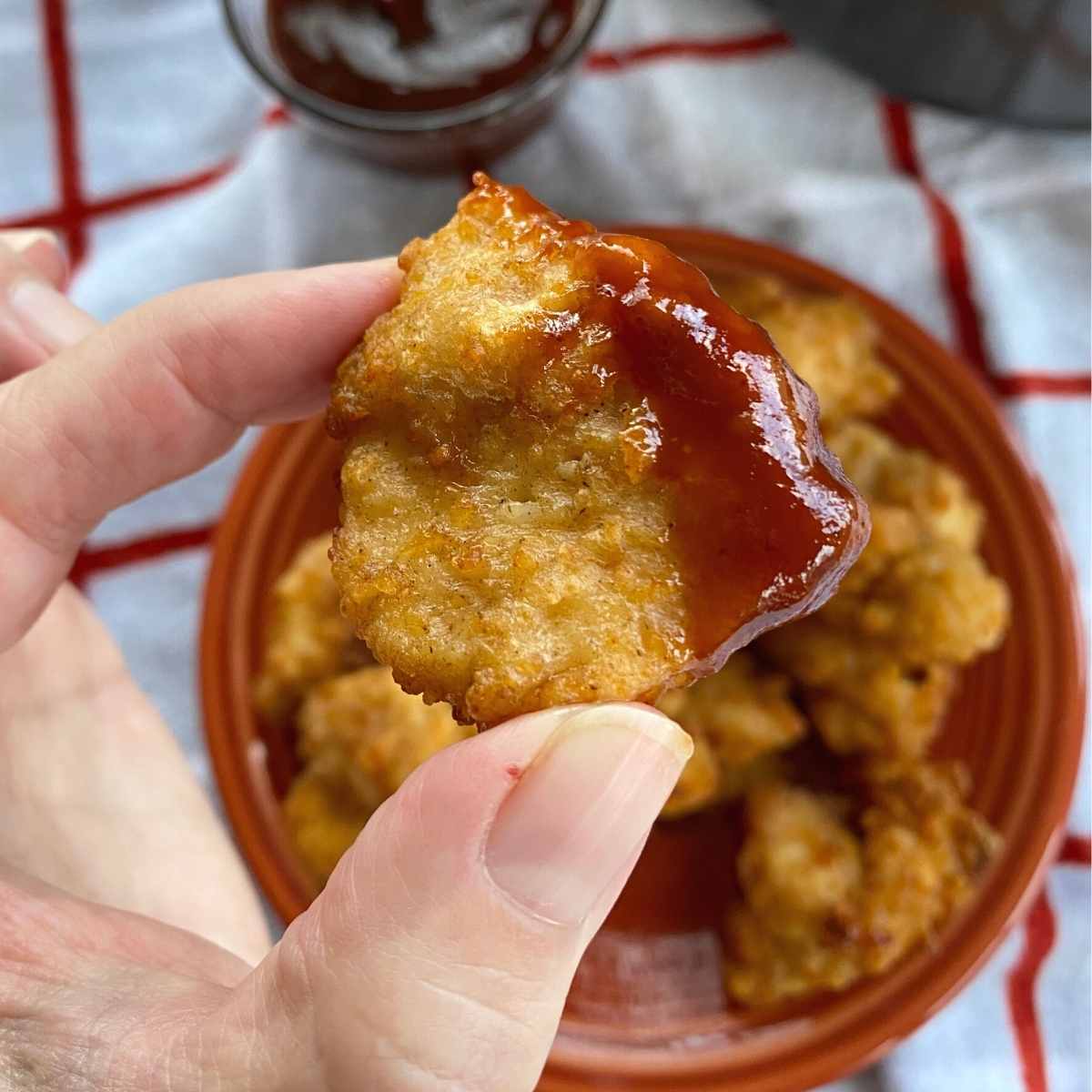Fingers holding a chicken nugget cooked in an air fryer dipped in BBQ sauce.