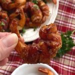 Fingers holding a stuffed bacon wrapped shrimp with more on a plate below.