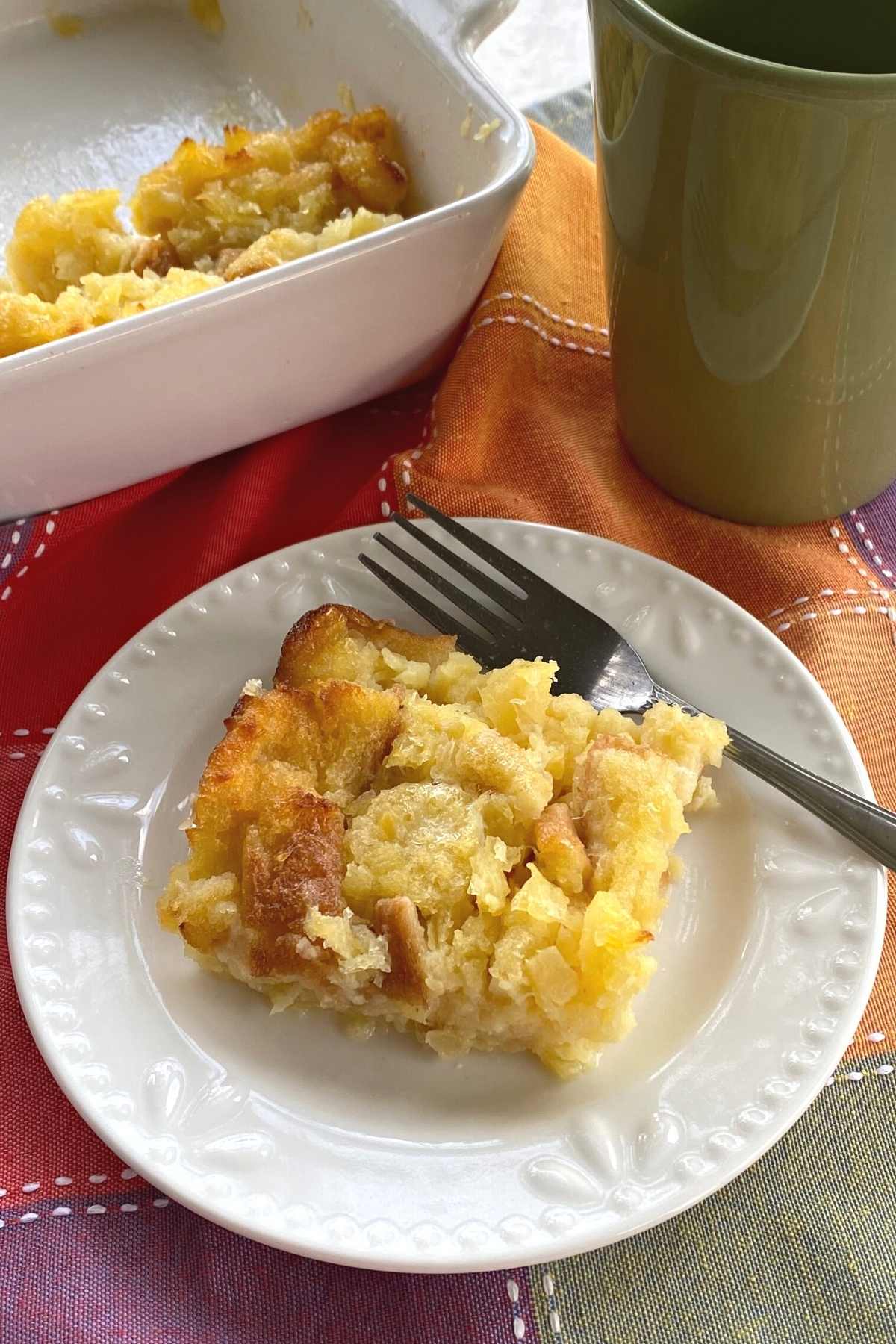 Serving of pineapple souffle on a plate with a baking dish and coffee cup behind it.