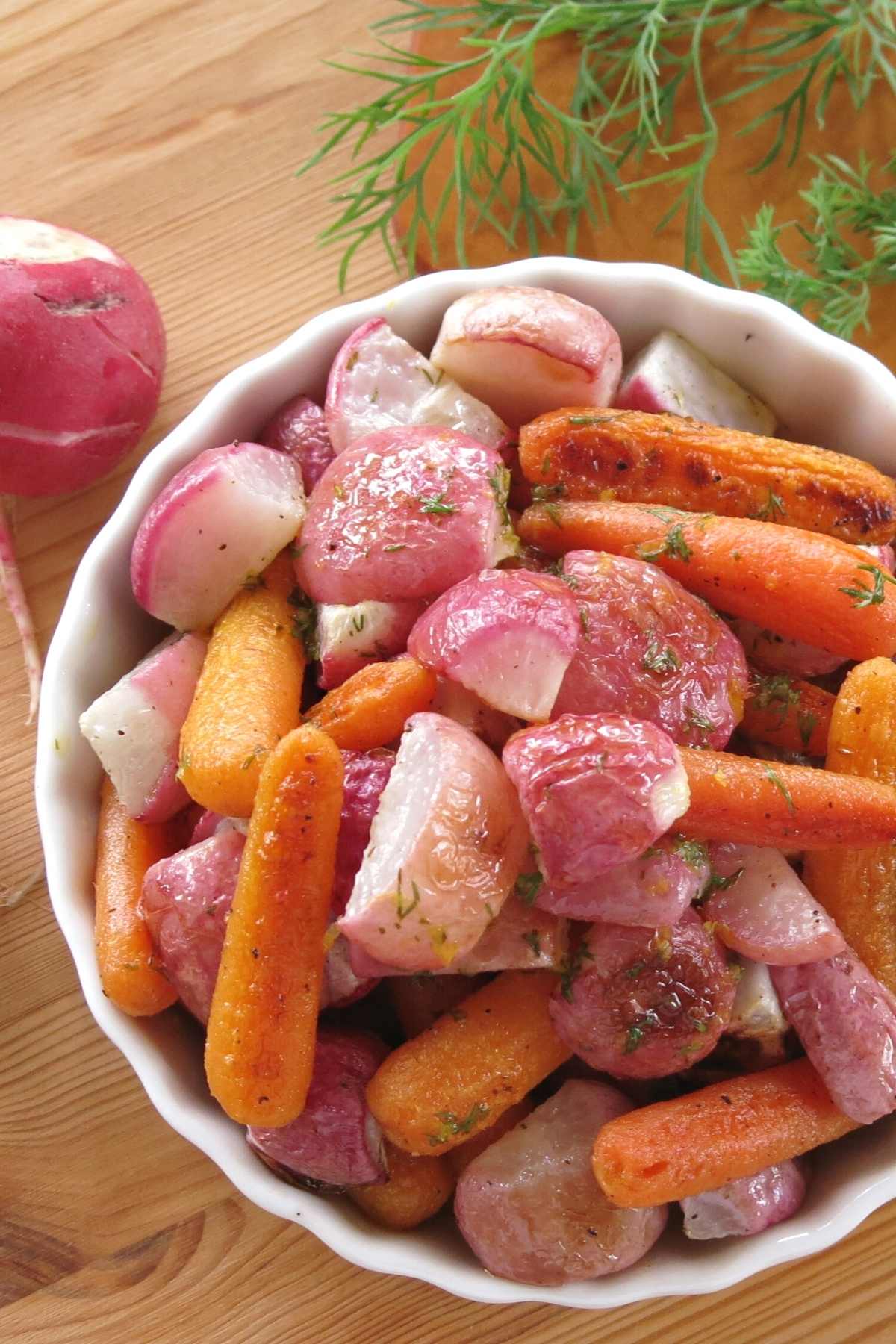 Oven roasted radishes and carrots in a serving dish with fresh dill in the background.