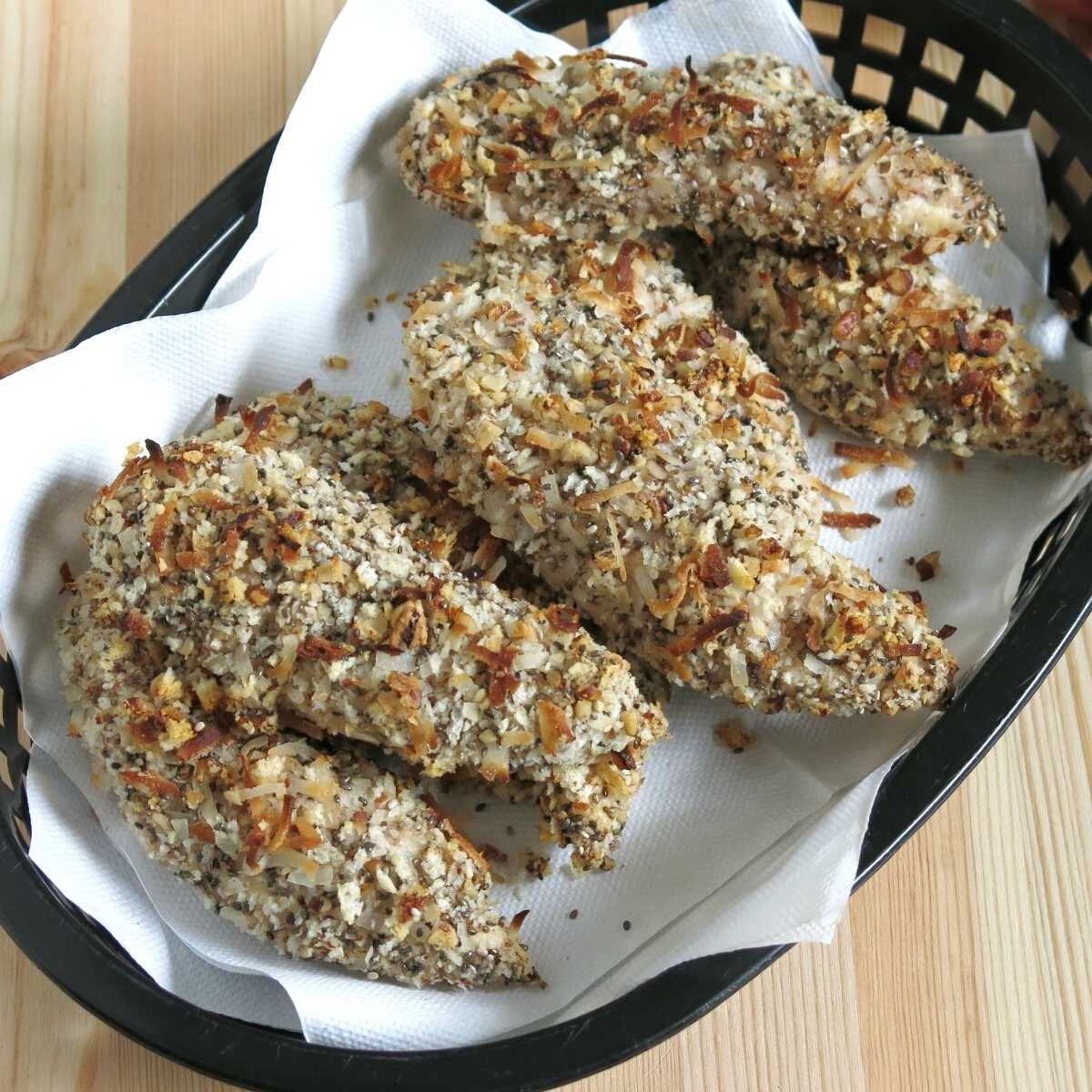 Easy baked chicken tenders breaded with healthy combination of panko, coconut, pecans, and chia seeds in a basket lined with a napkin.