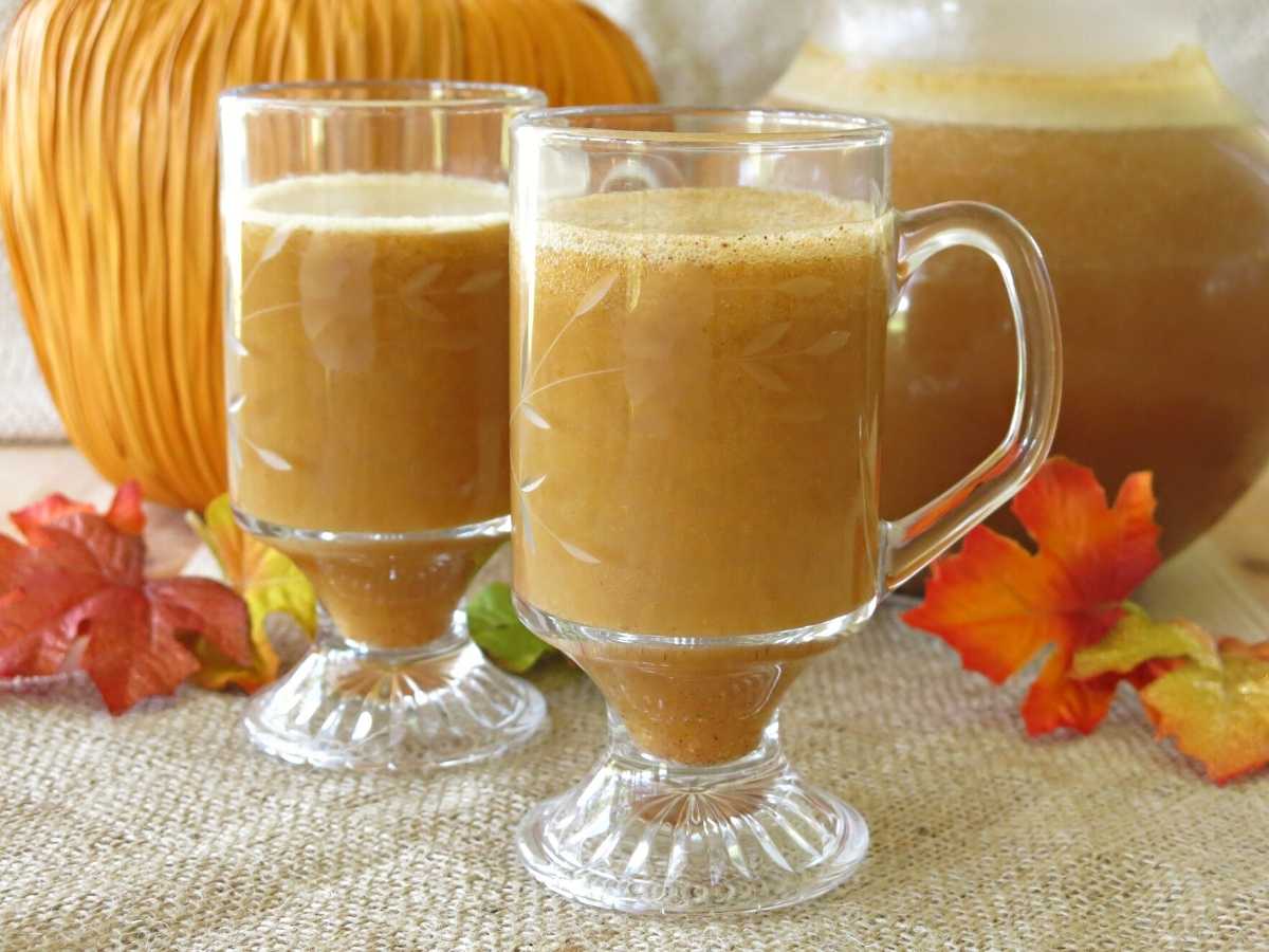 2 mugs of pumpkin juice with a large pitcher and pumpkin behind it.