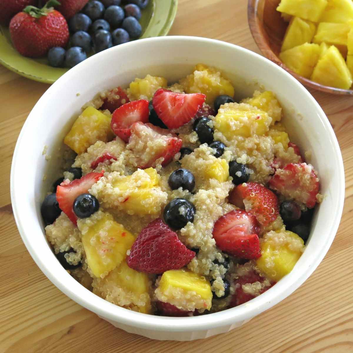 Quinoa fruit salad with honey lime dressing in a serving bowl with strawberries, blueberries, and pineapple in bowls around it.