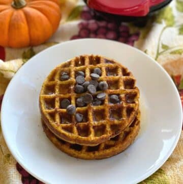 Two pumpkin chaffles topped with chocolate chips stacked on a plate with a pumpkin and waffle maker behind them.
