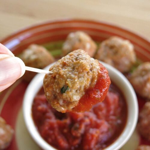 Cheesy baked chicken parmesan meatball dipped in marinara on a toothpick with a plate of more meatballs below it.