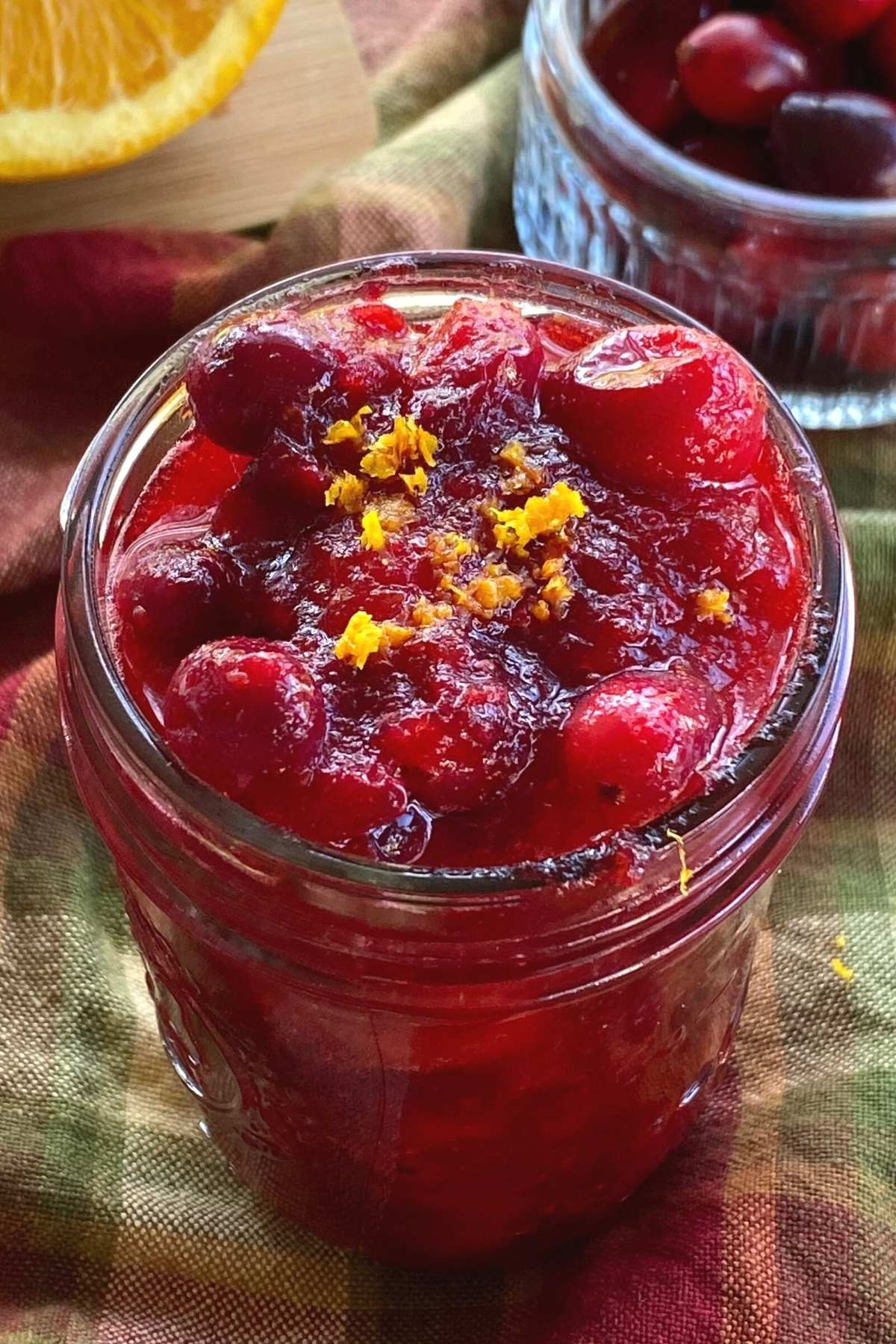 Whole cranberry sauce with less sugar in a mason jar with cranberries and an orange behind it.
